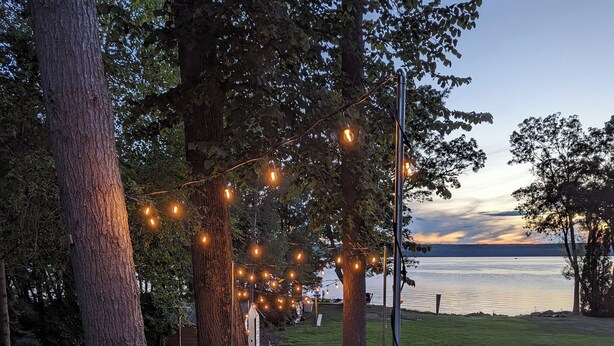 A stone pathway lined with trees and string lights leading to a body of water.