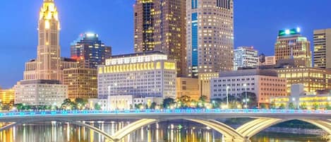 A city skyline with illuminated buildings and a bridge reflecting in the water.