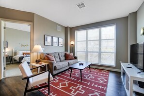 A modern living room with a sofa, coffee table, and TV stand. There is a window with blinds, a lamp, and a patterned rug. - Downtown Designer 2BR Parking W D Gym Pool (Columbus)
