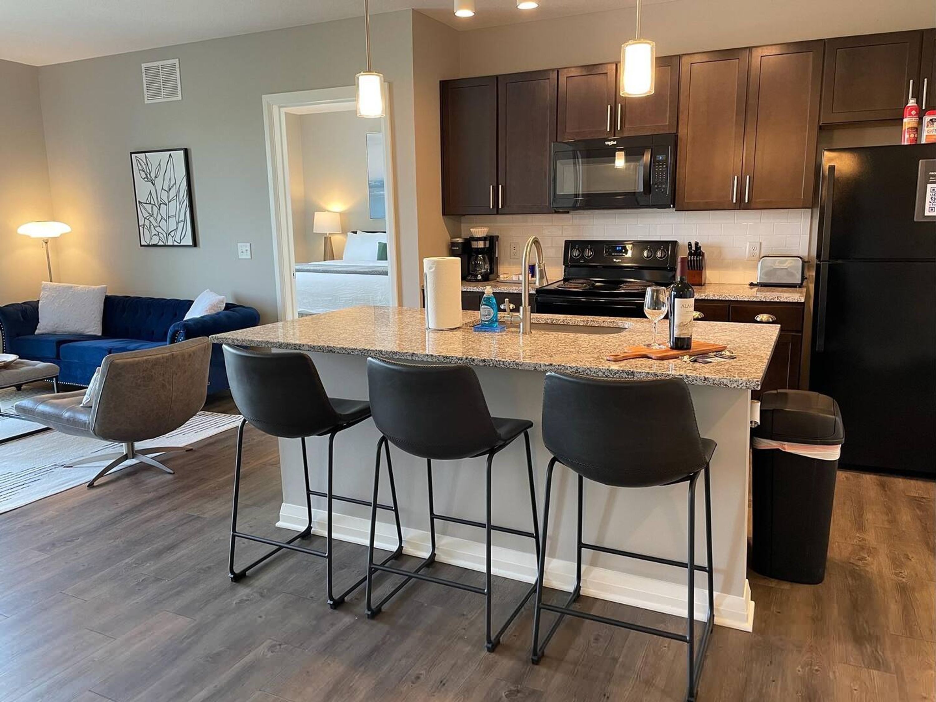 A modern kitchen with dark wooden cabinets, a stainless steel refrigerator, a microwave, and a gas stove. There are four black bar stools around the island with a granite countertop.