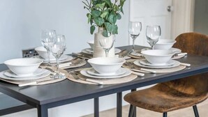 Una mesa de comedor preparada con platos, cuencos y cubiertos blancos, junto a un florero con follaje verde.