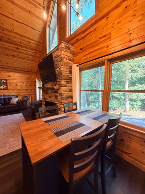 A wooden cabin interior with a stone fireplace, dining table, chairs, and large windows.