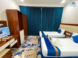 A hotel room with two beds, a desk with a computer, and a large window with blue curtains. - Zad Al-Mashaer Hotel (Makkah)