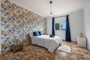 Un dormitorio con cama, mesita de noche, lámpara, ventana con cortinas y un papel tapiz con motivos de hojas tropicales.