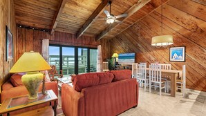 A cozy living room with a wooden ceiling, a ceiling fan, a dining area with a table and chairs, and a large window offering a view. - Sea Dunes Green Turtle A3 (New Smyrna Beach)