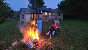 A person sitting by a campfire in a grassy yard with a house in the background. - House of Friendship, Connect in a Relaxing Getaway near Red River Gorge (Wellington)