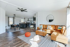Ein großzügiger Wohnbereich mit einer Couch, einem Hocker und einem Esstisch mit Stühlen. Im Hintergrund ist eine Küche und ein Deckenventilator zu sehen.