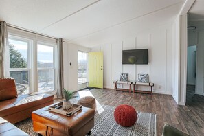 A modern living room with a sofa, ottoman, and a red pouf, a yellow door, and a TV mounted on the wall.