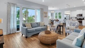 A spacious living room with a grey sofa, a round woven ottoman, and a dining area with patterned chairs.