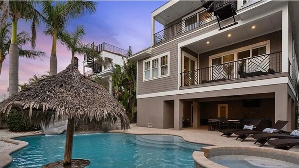 A modern two-story building with a swimming pool, a thatched-roof structure, and lounge chairs.