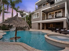A modern two-story building with a swimming pool, a thatched-roof structure, and lounge chairs. - Pool with with water slide & grotto! Sleeps 14! Walk to Beach! (Holmes Beach)