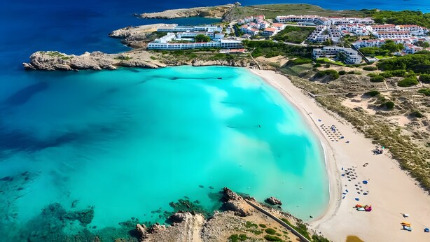 Oversigtsbillede af et kystresort med en strand, klart turkisblåt vand og en gruppe bygninger.