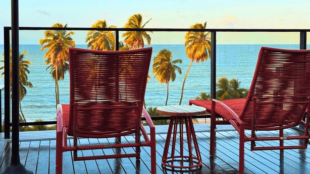 Ein Balkon mit zwei roten Korbstühlen und einem kleinen Tisch, mit Blick auf einen Strand mit Palmen und dem Meer.