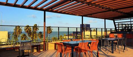 An outdoor terrace with tables and chairs, overlooking a beach and ocean.