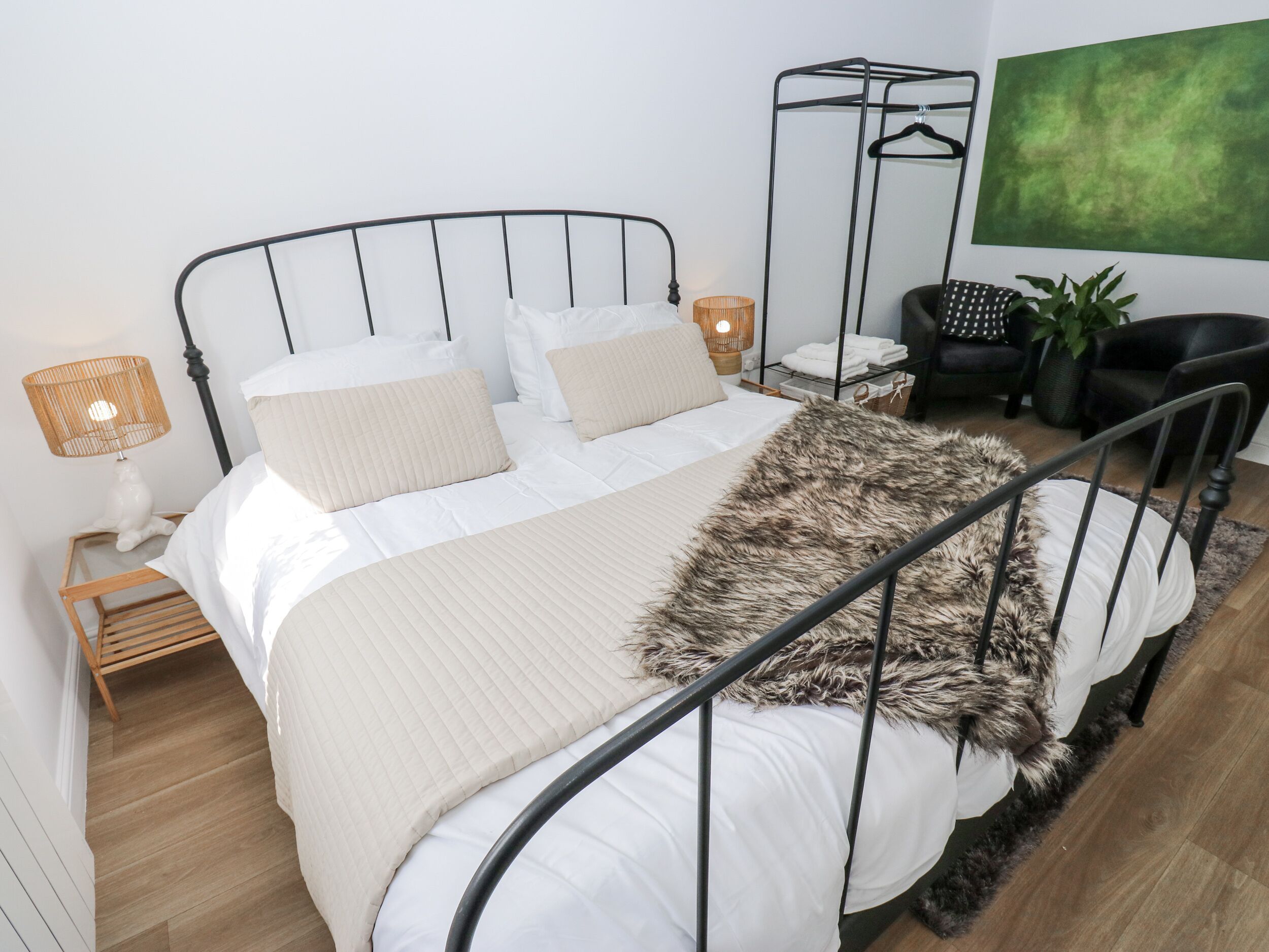 A neatly arranged bedroom with a metal bed frame, white bedding, a fur throw, a bedside lamp, and a green abstract painting on the wall.