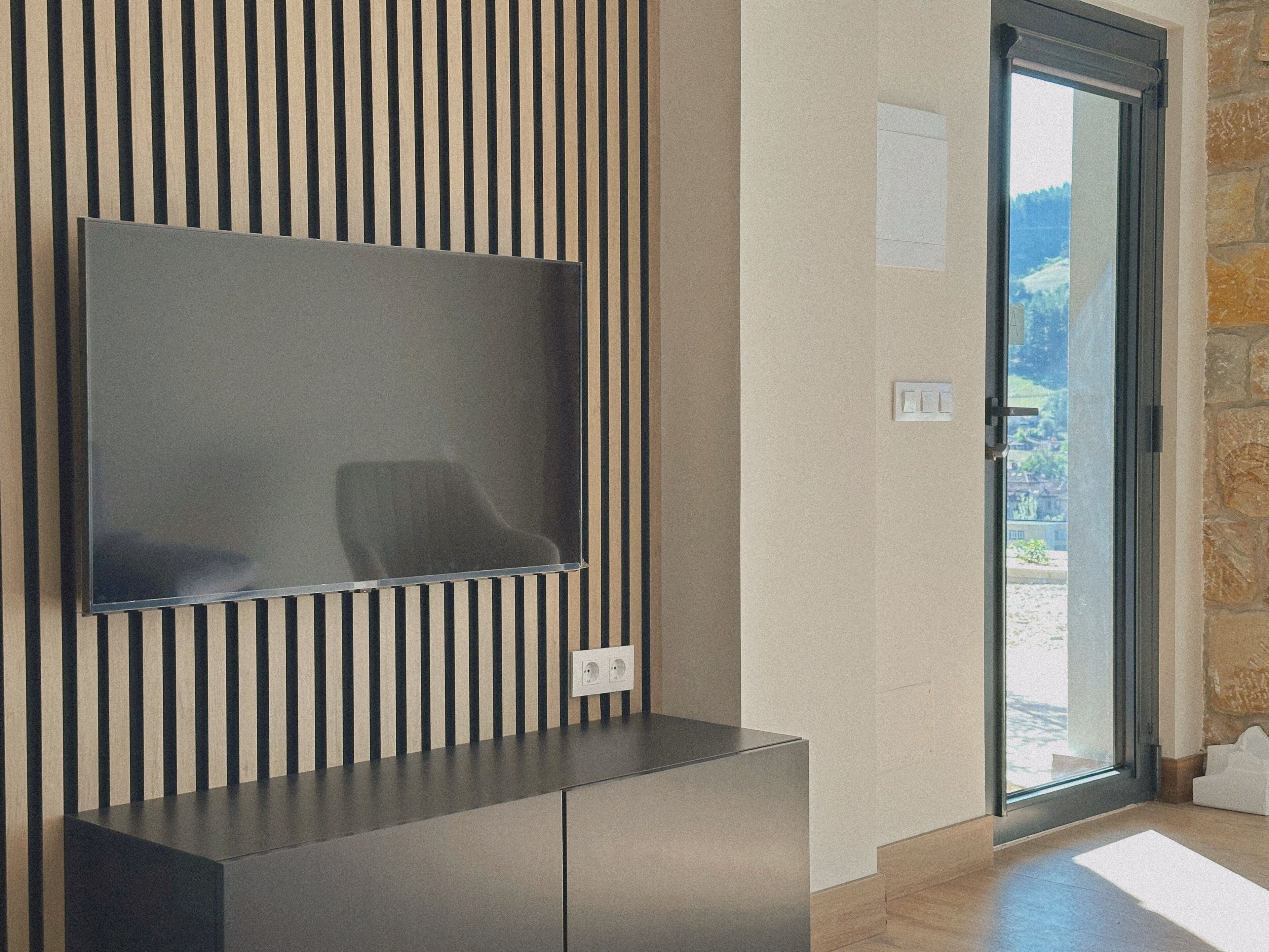Un interior moderno con un televisor de pantalla plana montado en una pared a rayas verticales, una puerta que da a un área exterior y una pared de piedra.