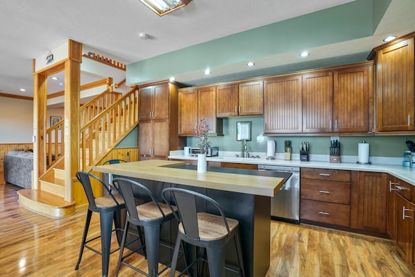 A modern kitchen with wooden cabinets, a central island with bar stools, a built-in dishwasher, and a staircase leading to another level.
