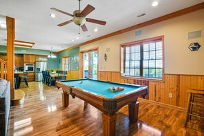 A pool table in a room with wooden floors and walls, a ceiling fan, and a kitchen area visible through an open door.