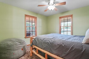 Une chambre à coucher avec un lit, un ventilateur de plafond et deux fenêtres équipées de stores en bois.