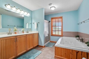 Une salle de bain avec des meubles en bois, un grand miroir, une baignoire et une fenêtre avec des stores.