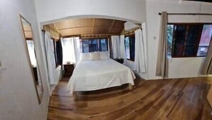Un dormitorio con piso de madera, una cama con colcha blanca y un ventanal con cortinas.