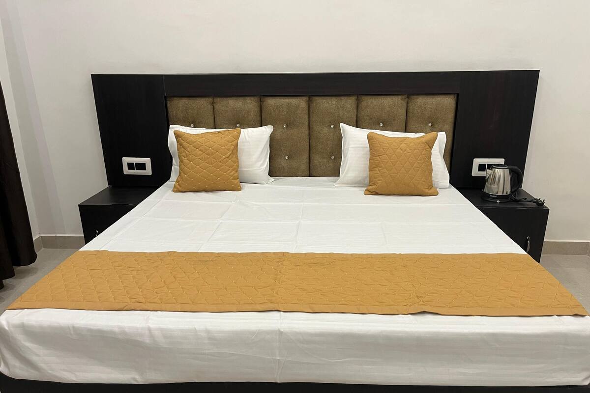 Una cama bien hecha con una colcha blanca y marrón, dos almohadas blancas y un plaid marrón acolchado. Hay dos mesitas de noche, una con una tetera.