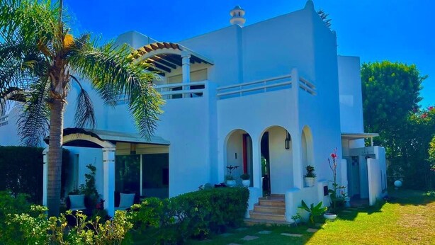 A white building with a covered porch, arched doorways, and a small staircase leading to the entrance. - Villa au calme, jardin spacieux et confort moderne (Skhirat)