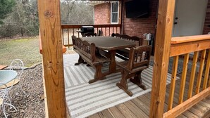 Una terraza techada al aire libre con una mesa y sillas de madera, una parrilla y un televisor montado en la pared.