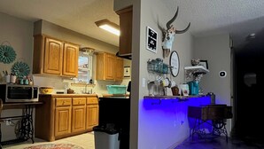 Una cocina con gabinetes de madera, un microondas y un lavamanos. Hay un bote de basura y un plato para mascotas en el suelo.