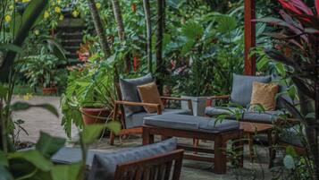 Un área de estar al aire libre con mobiliario de madera y una mesa, rodeada de abundante follaje y plantas.