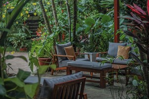 Un área de estar al aire libre con mobiliario de madera y una mesa, rodeada de abundante follaje y plantas.