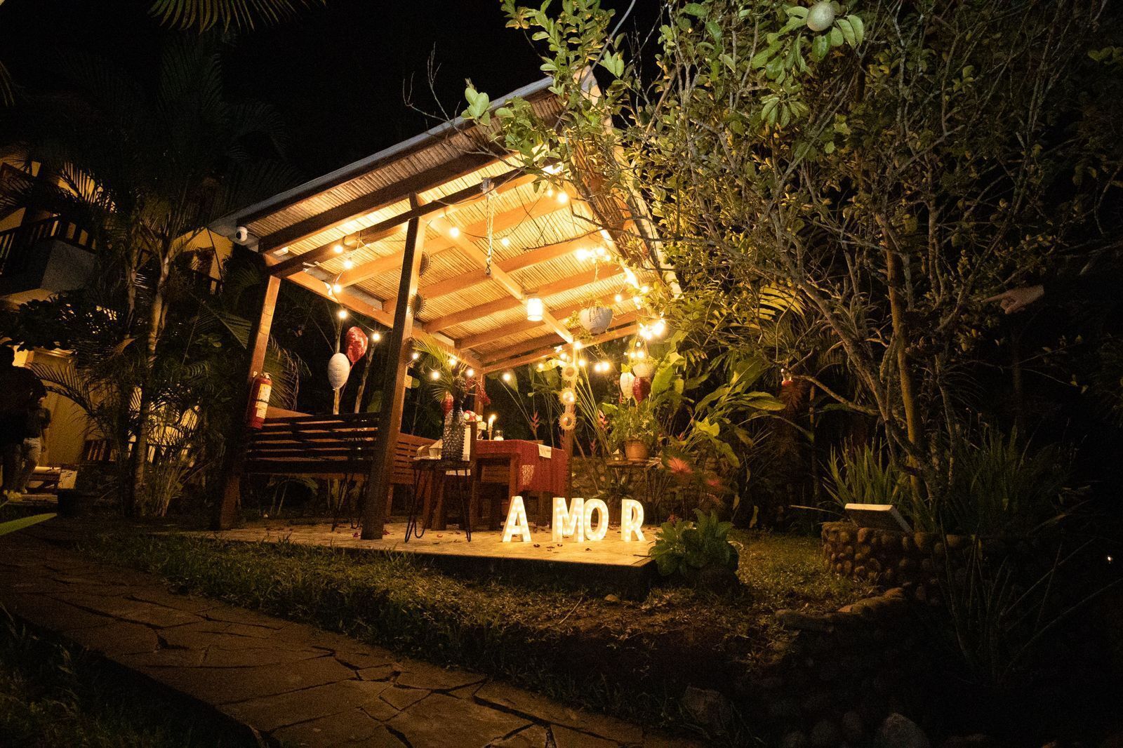 An outdoor dining area with a covered seating arrangement, surrounded by lush greenery and a well-lit ambiance.