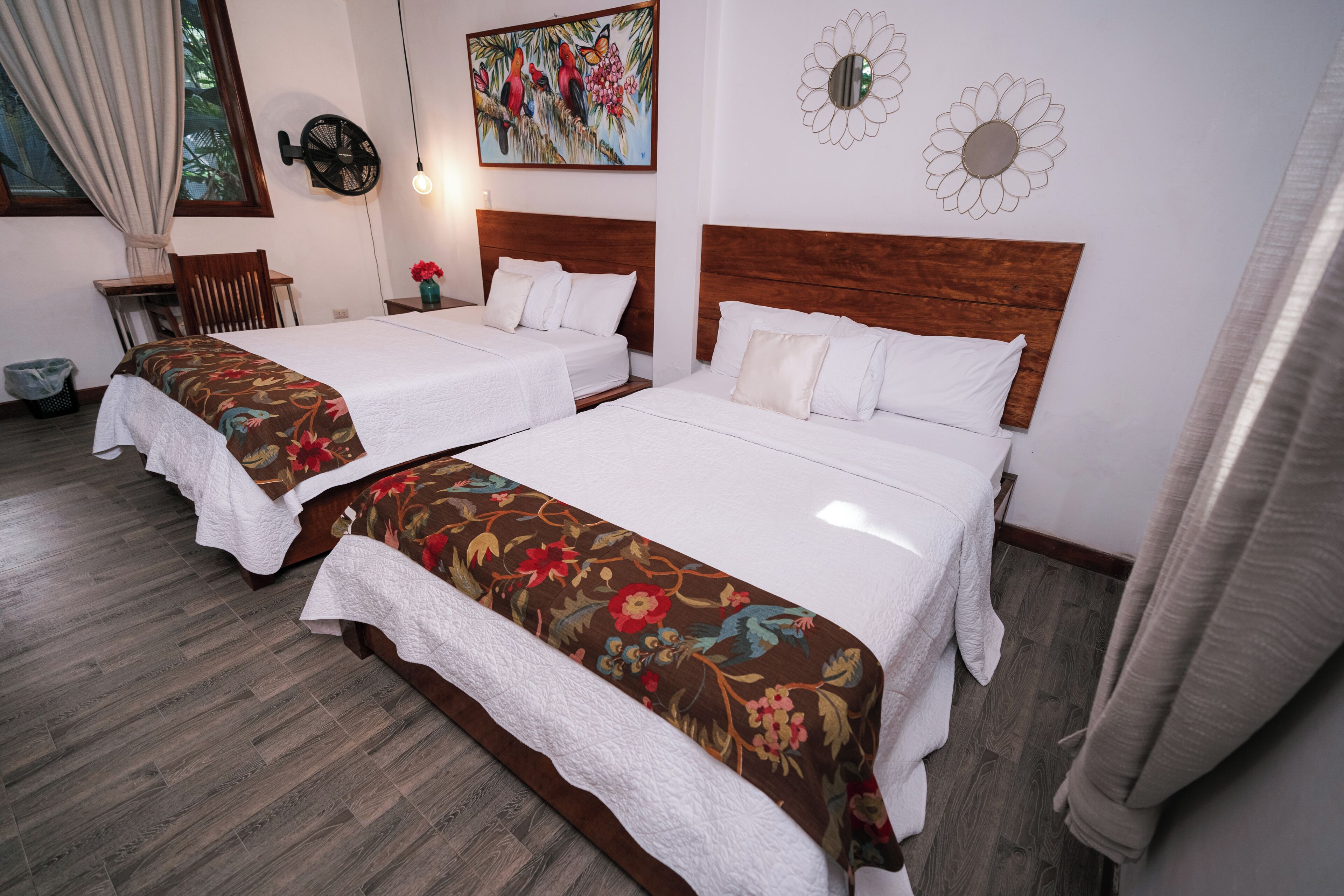 A hotel room with two beds, a wooden headboard, a desk, a chair, and a window with curtains.