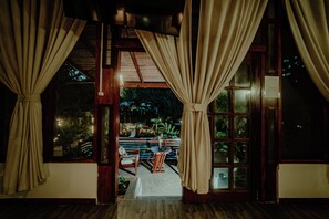 A room with wooden floors, a window with curtains, and a view of an outdoor seating area with tables and chairs. - Villa Lu Amazon Lodge (Tarapoto)