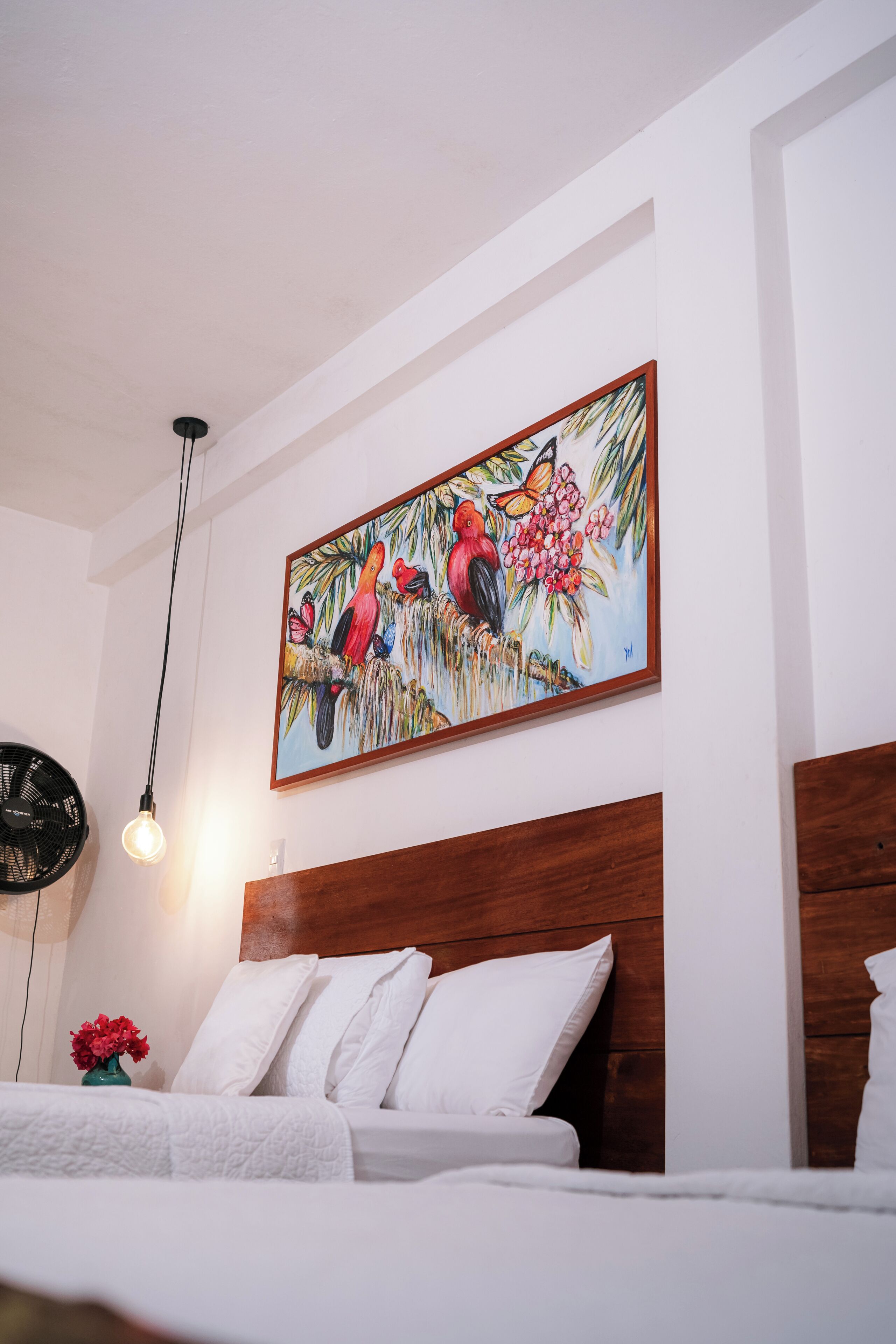A hotel room with a wooden headboard, white bedding, a wall-mounted painting of tropical birds, and a small table with a vase of flowers.