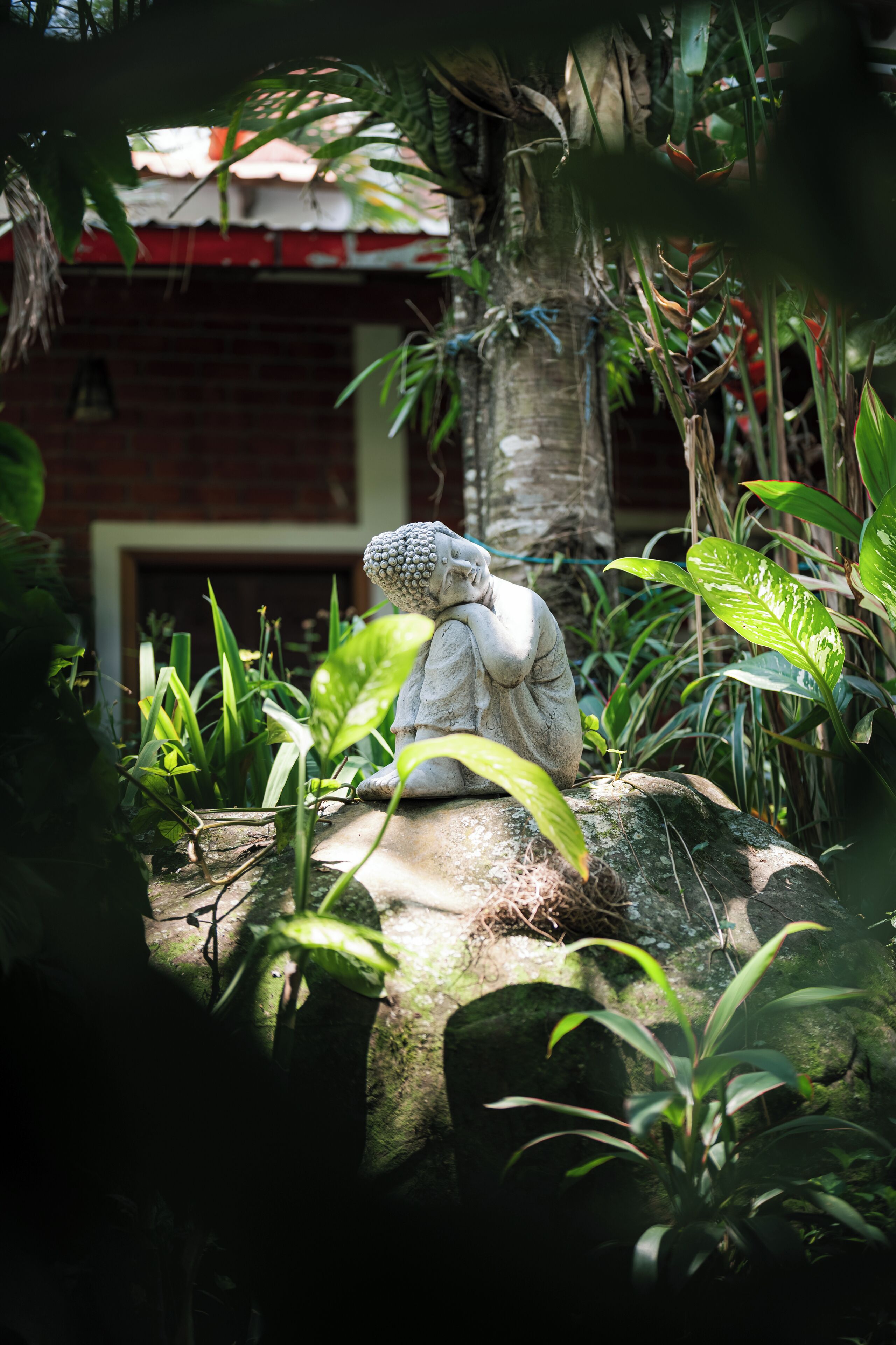 A statue of a seated figure in a garden with lush greenery and a brick building in the background.