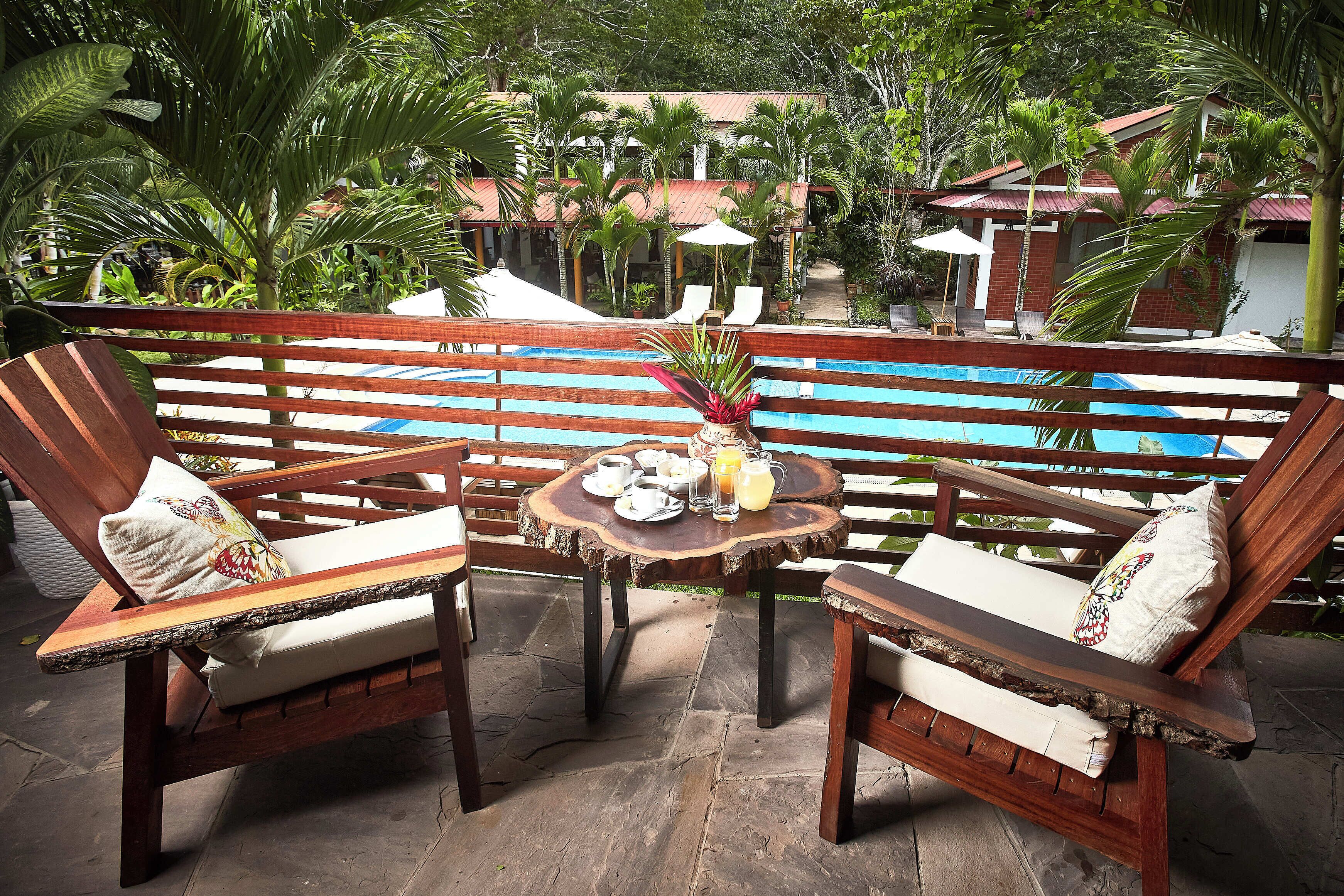 A wooden deck with two lounge chairs, a small round table with a vase and a glass of juice, and a view of a pool and a building in the background.