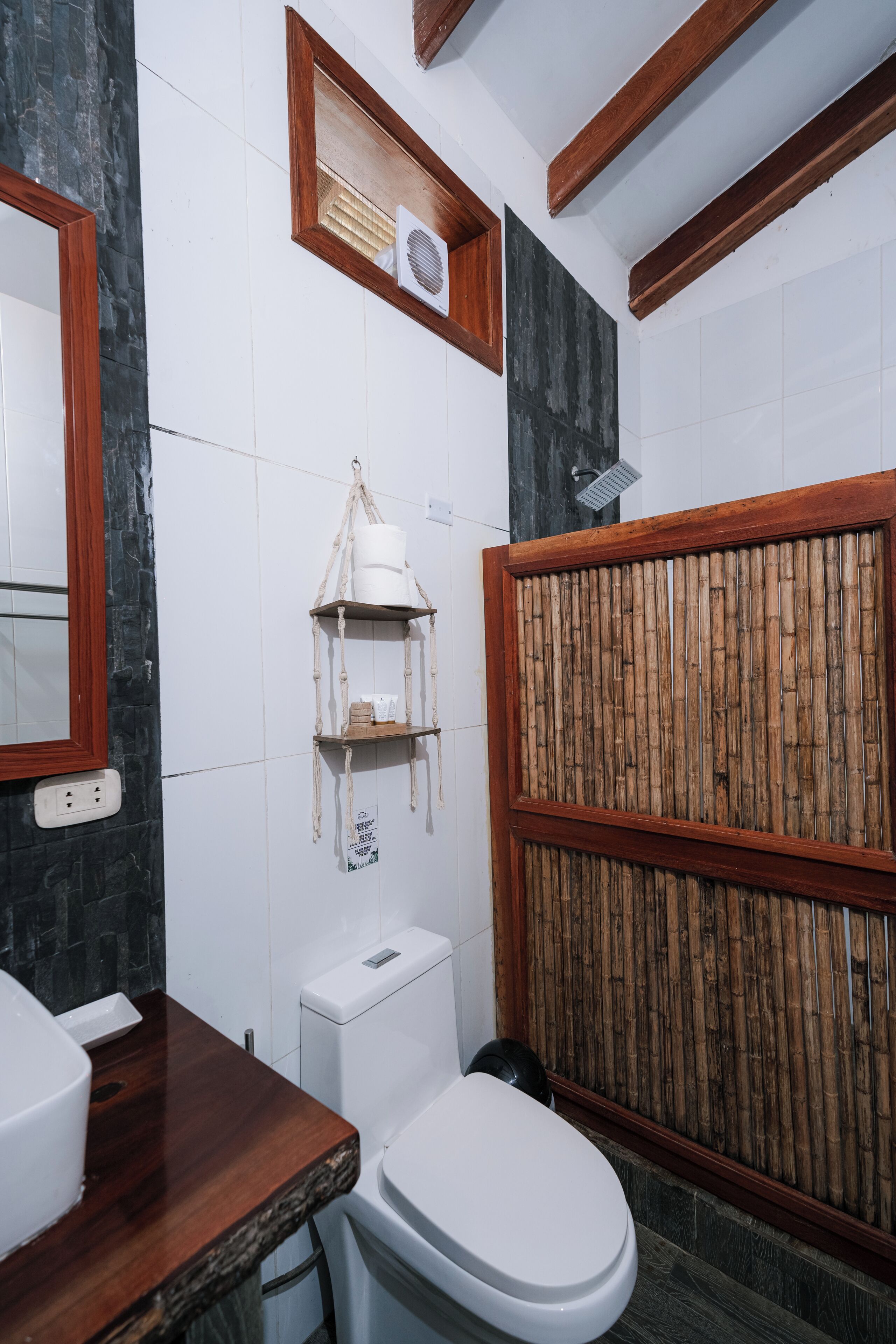 Ein Badezimmer mit einer weißen Toilette, einem hölzernen Waschtisch, einer Duschkabine aus Bambus und einem Wandregal mit Toilettenartikeln.