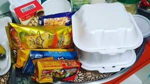 Verschiedene Speisen und Behälter auf einer Tablett, darunter verpackte Snacks, eine Plastikbox und ein Plastikbecher.