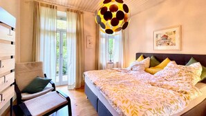 Une chambre à coucher avec un grand lit, une chaise, un luminaire suspendu et une fenêtre avec des rideaux.