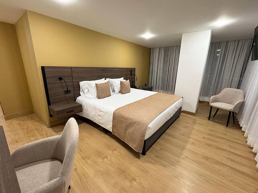 A hotel room with a large bed, a bedside table, a chair, and a view of the outside through curtains.
