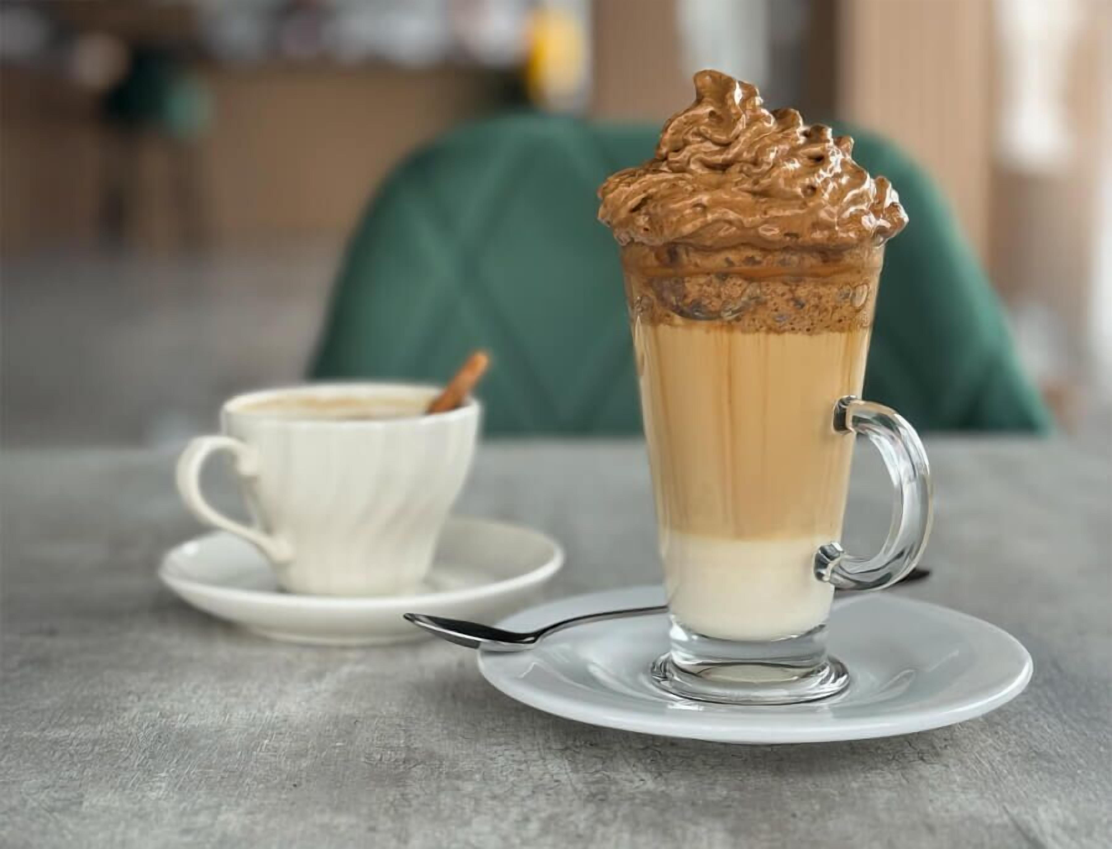 Ein hohes Glas mit einem geschichteten Getränk und Schlagsahne, zusammen mit einer kleinen Tasse und einem Untertasse auf einem Tisch.