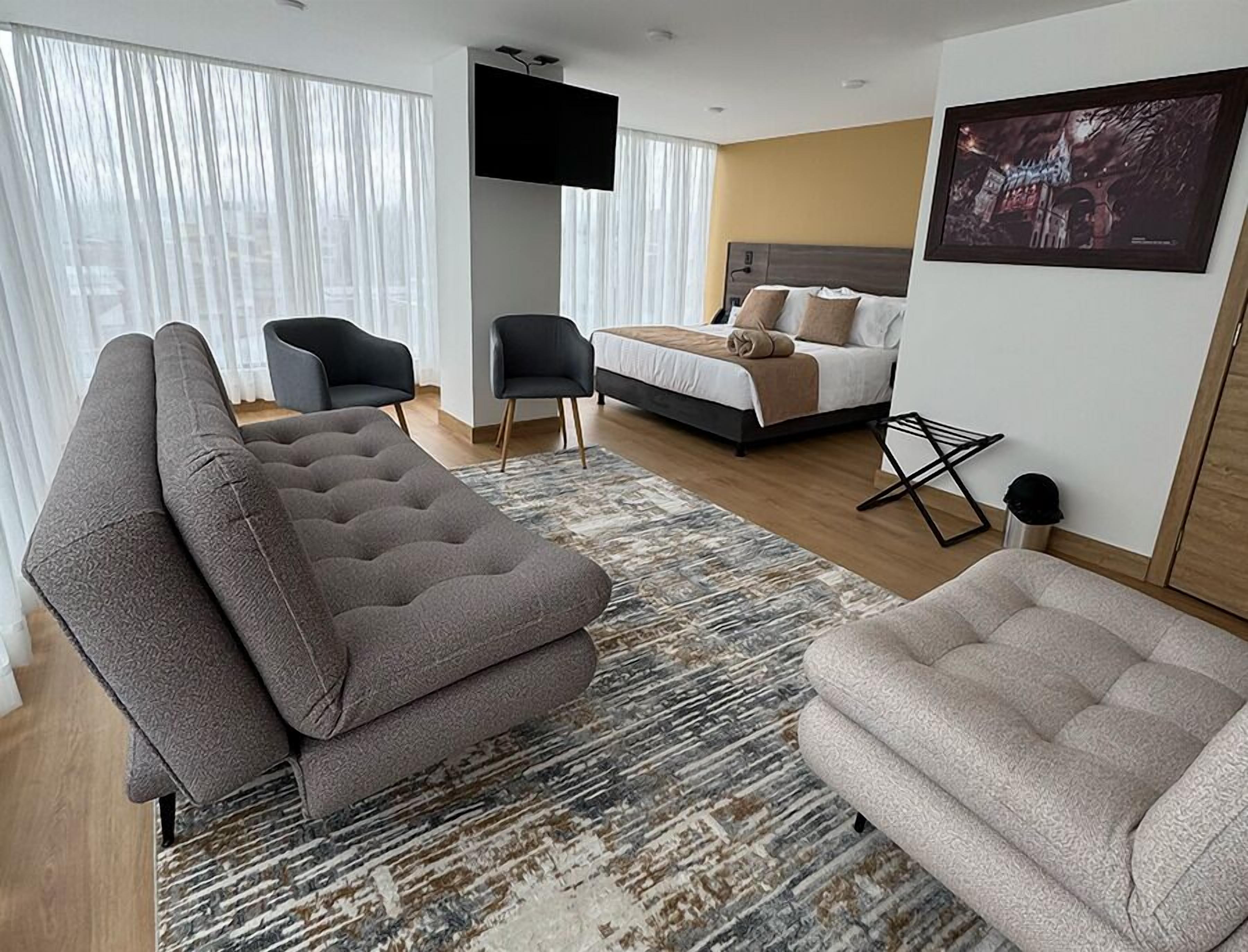 Une chambre d’hôtel moderne dotée d’un grand canapé d’angle, d’un lit simple, d’un téléviseur à écran plat et d’une grande fenêtre avec des rideaux légers.