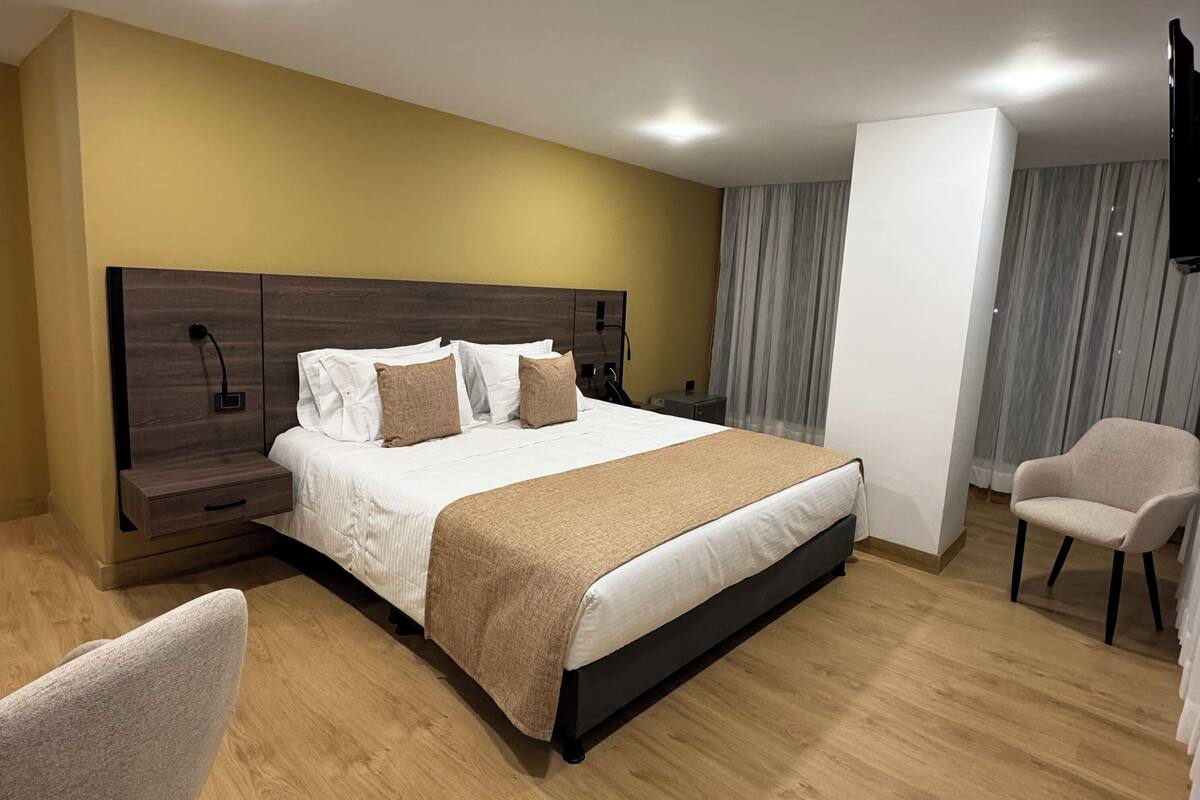 Quarto de hotel moderno com uma cama grande, mesa de cabeceira, uma cadeira e televisão de tela plana na parede.