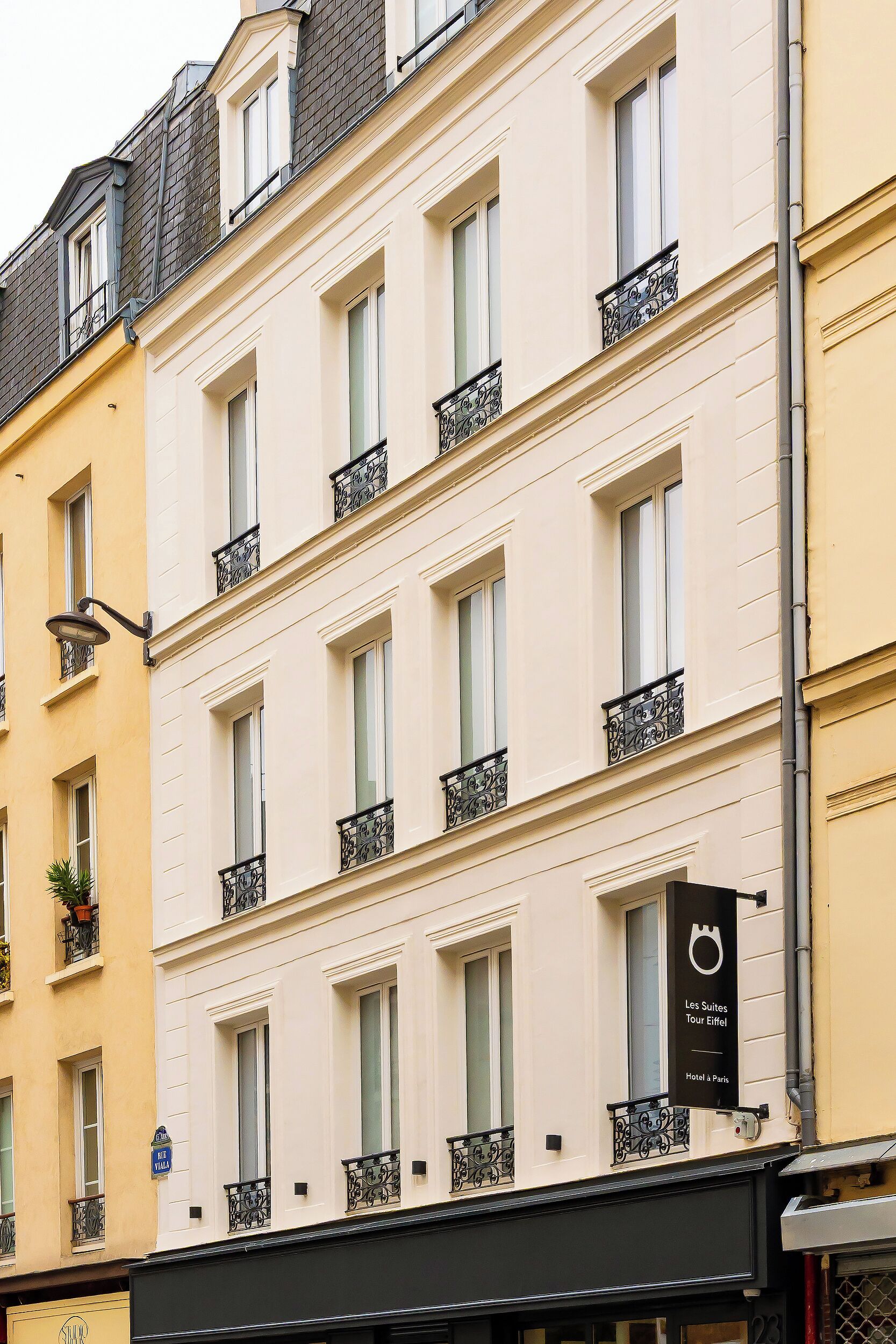 A multi-story building with a light-colored facade, multiple windows with black wrought iron balconies, and a sign indicating it is a hotel.