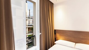 Habitación de hotel con cama, cabecero de madera, ventana con cortinas y vista a un paisaje urbano que incluye una torre.