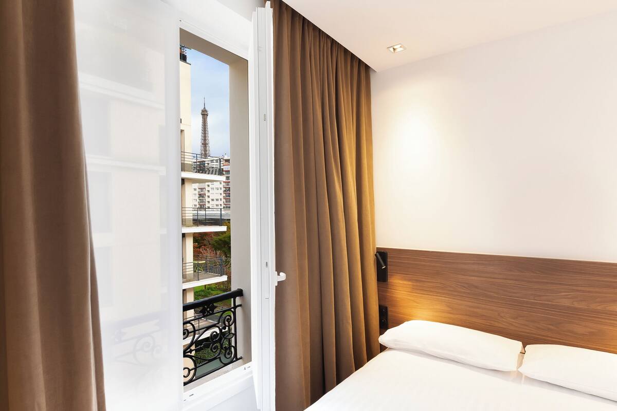 Ein Hotelzimmer mit einem Bett, einem Holzkopfende, einem Fenster mit Vorhängen und Blick auf eine Stadtlandschaft mit einem Turm.