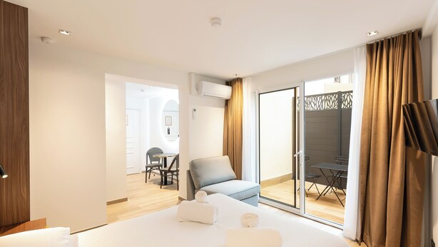 Habitación de hotel con una cama grande, una zona de estar con una silla y una mesa, y un ventanal corredero que da a un patio exterior.