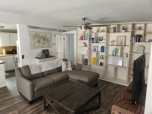 Ein Wohnzimmer mit einem Ecksofa, einem Couchtisch, einem Fernseher, einem Bücherregal und einem Deckenventilator.