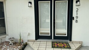 A house with a black front door, two windows with blinds, a small garden with a stone lantern, and a number '2198' on the wall.